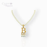 Gold necklace with 'B' pendant and pearl drops on a white background, branded 'Crowns & Regalia'.